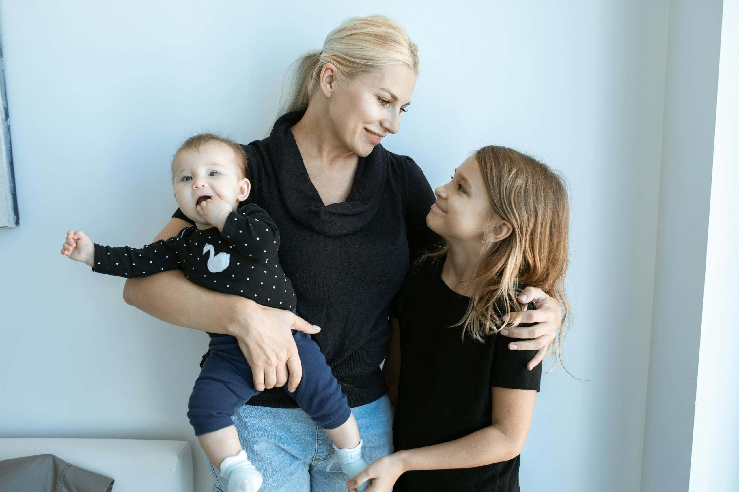 Happy mother holding her toddler and embracing her older daughter indoors.